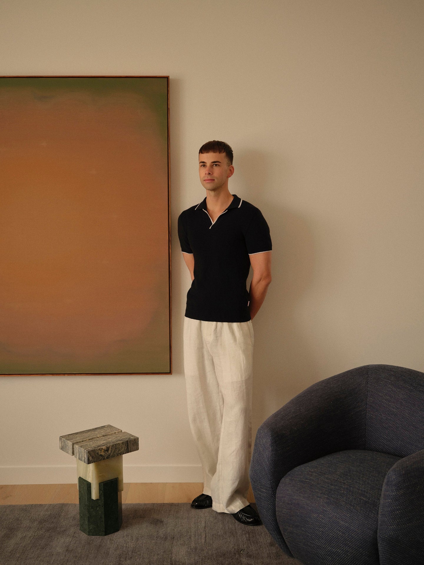 Man standing in a room with a large abstract painting on the wall, a gray chair, and a small stool.