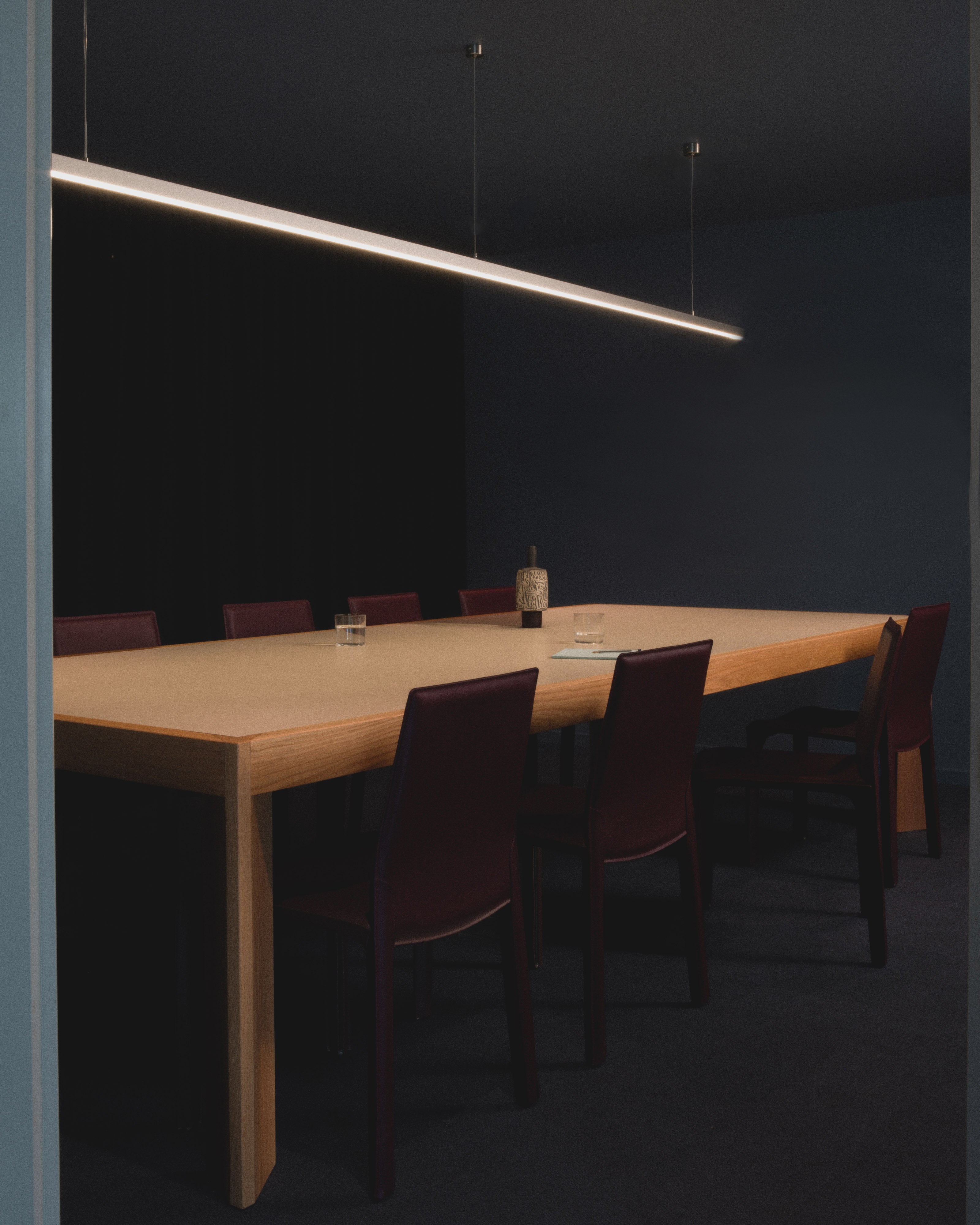 Moody dark commercial boardroom with a natural oak conference table, burgundy leather chairs, and a suspended linear LED pendant light — commercial office and hospitality interior design by Studio Nicholas Noosa Queensland Australia