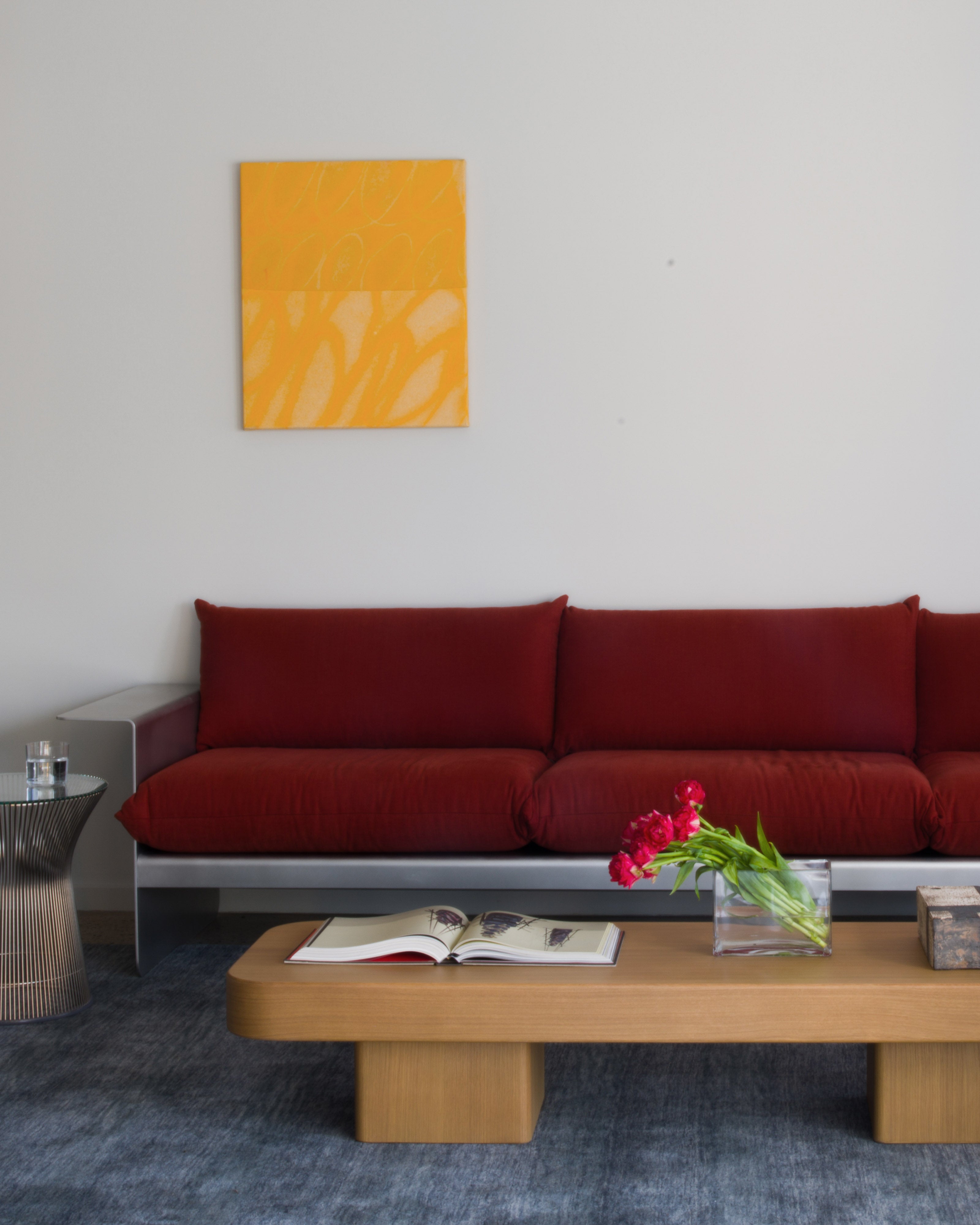 Residential living room with a red modular sofa beneath an orange abstract artwork, a low natural oak coffee table styled with an open book and red tulips, and blue-grey wool carpet — luxury residential interior design styling by Studio Nicholas Noosa Queensland Australia