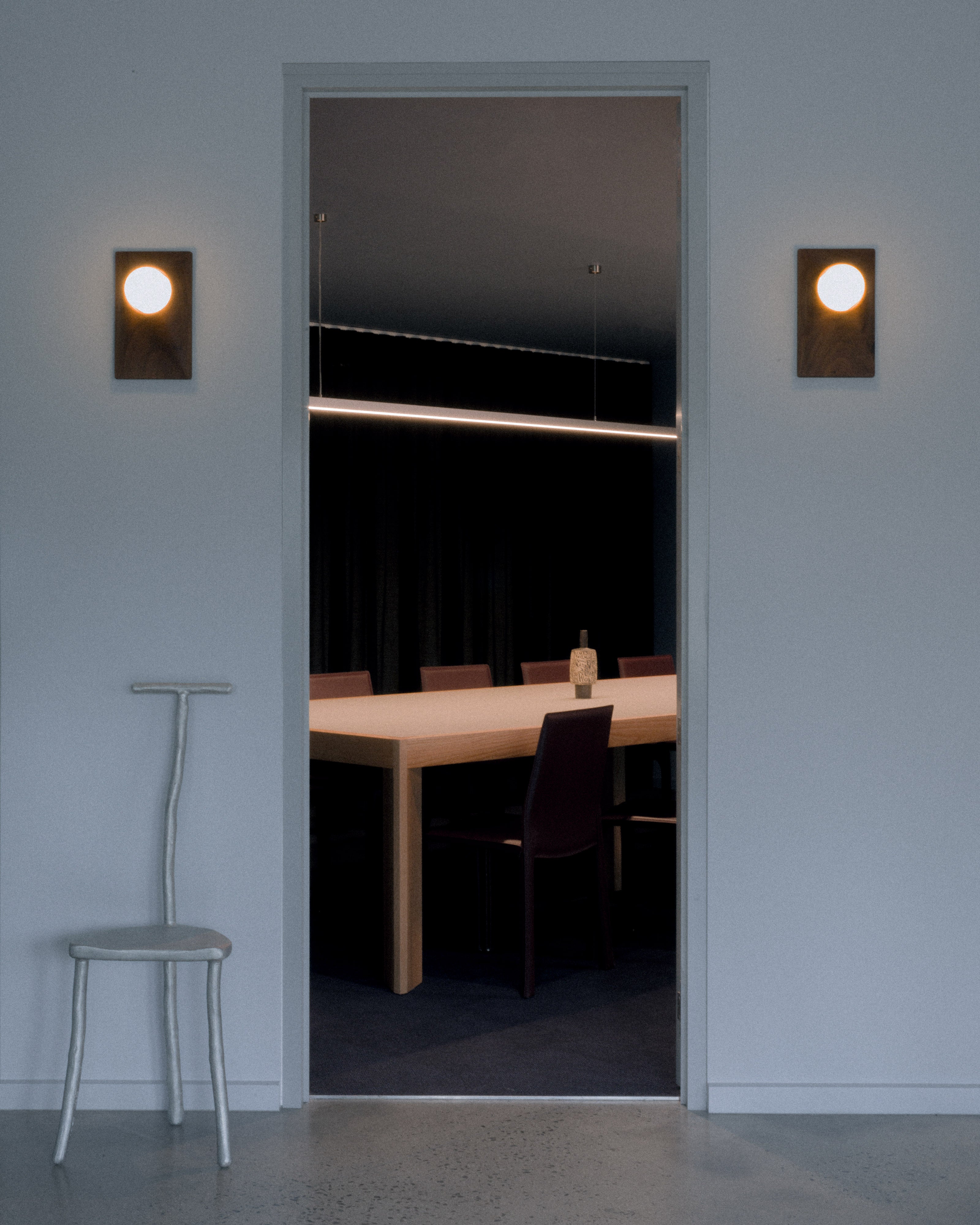 Commercial boardroom viewed through doorway with timber conference table, dark leather chairs, linear LED pendant lighting, and globe wall sconces flanking door opening on polished concrete floor — commercial and hospitality interior design by Studio Nicholas Noosa Queensland Australia