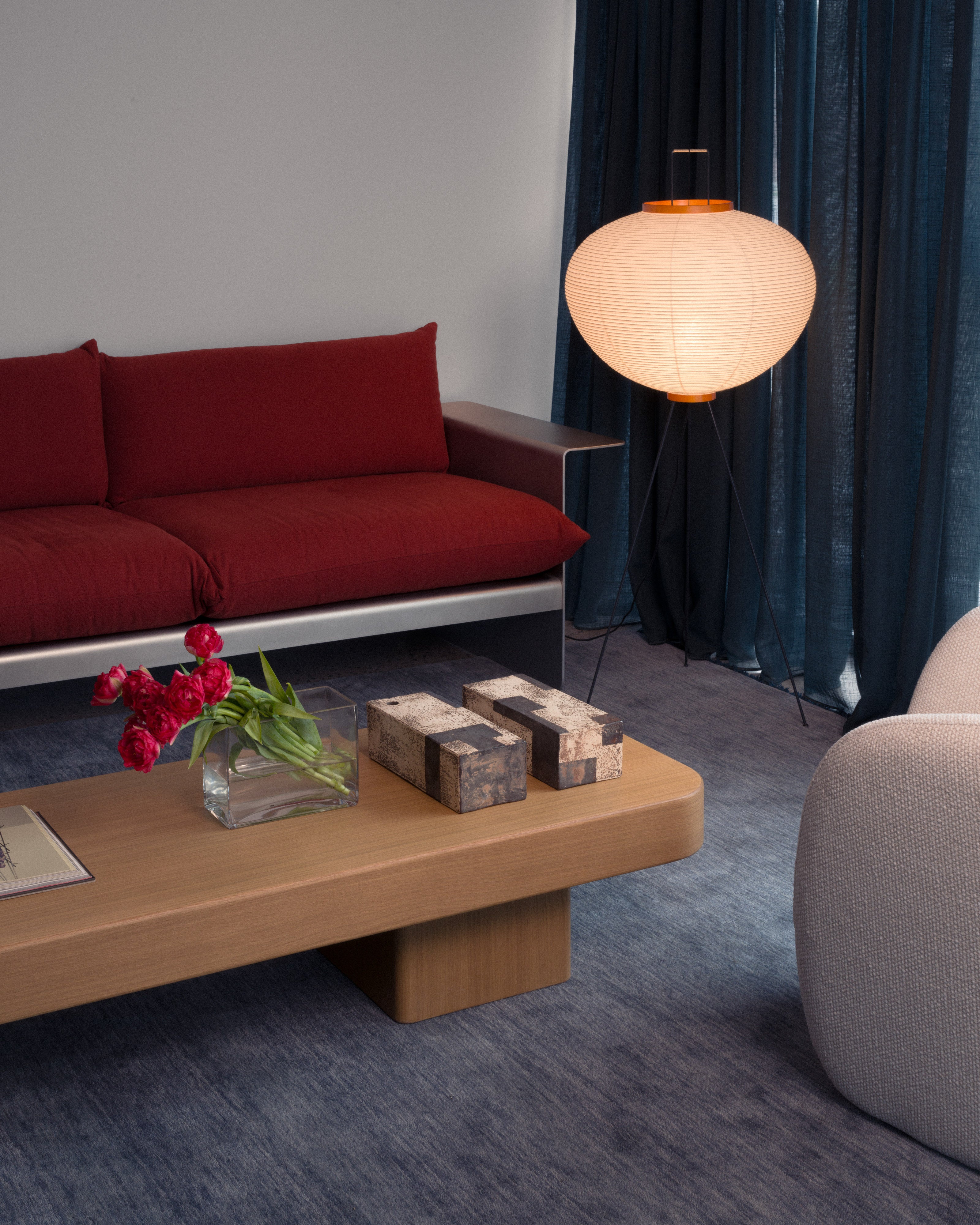 `Low natural oak coffee table styled with red tulips in glass vase and textured stone objects in a contemporary living room featuring a red upholstered sofa, Akari paper floor lamp, teal linen drape, and blue-grey wool carpet — luxury residential interior design by Studio Nicholas Noosa Queensland Australia`