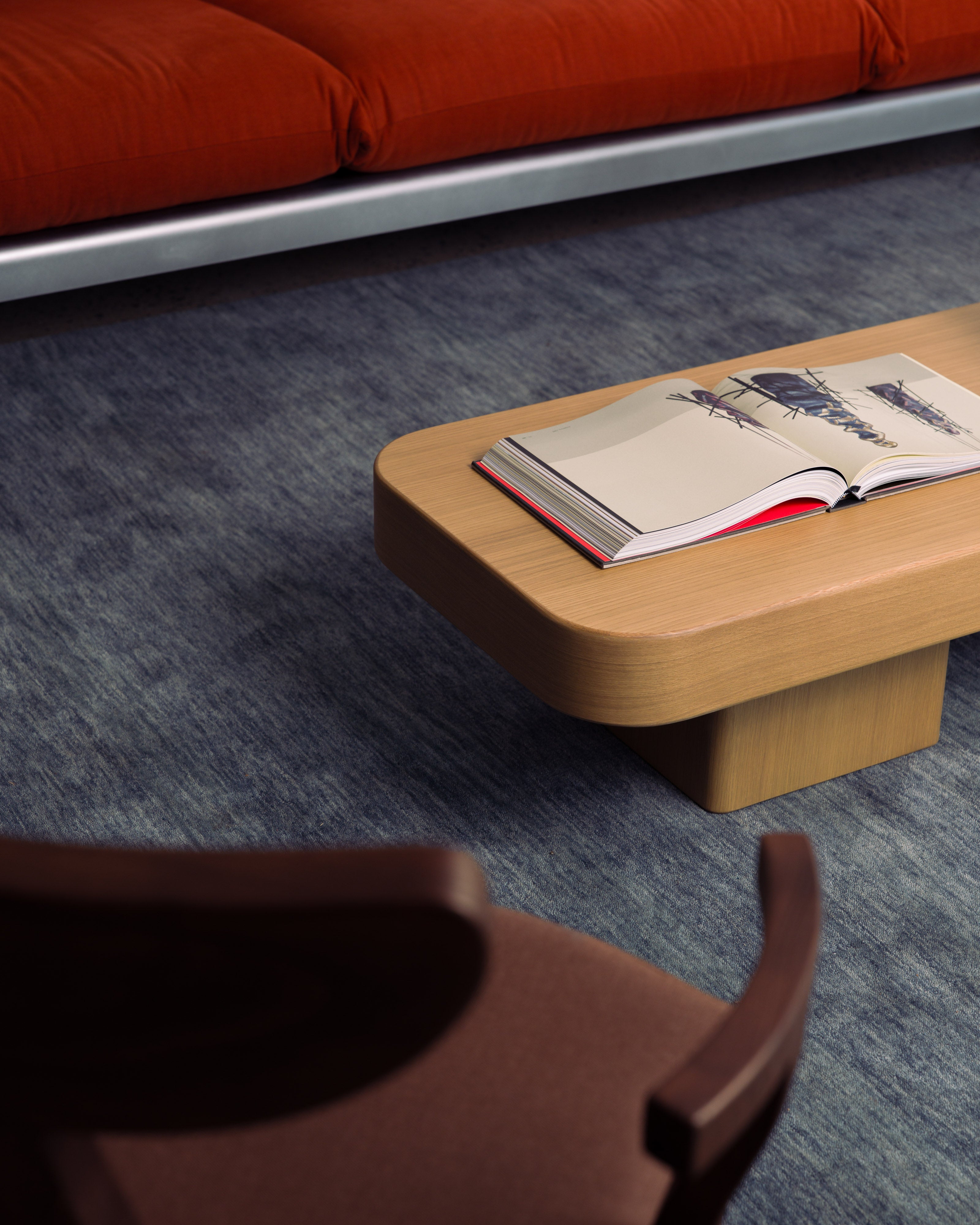 Low natural oak coffee table with an open book styled beside a red upholstered sofa and timber dining chair on blue-grey wool carpet — luxury residential interior design styling by Studio Nicholas Noosa Queensland Australia