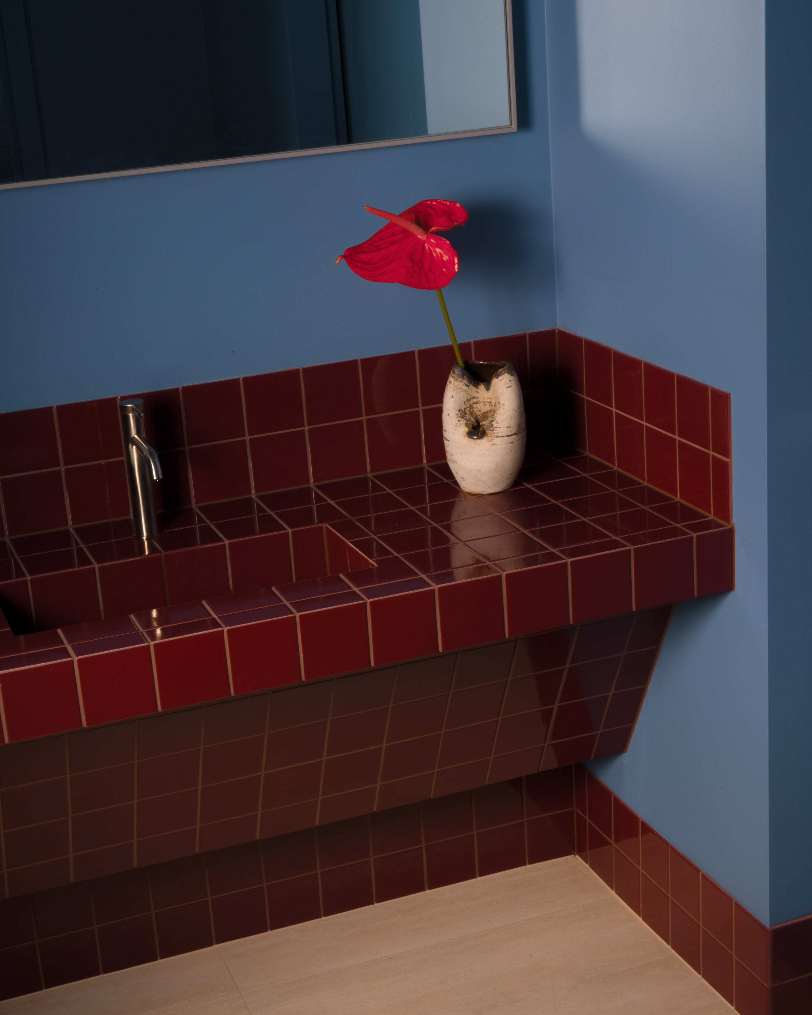 Deep burgundy tiled floating bathroom vanity with a stainless steel gooseneck tap, ceramic vase holding a red anthurium, blue-grey walls, wall mirror, and timber flooring — residential bathroom interior design by Studio Nicholas Noosa Queensland Australia