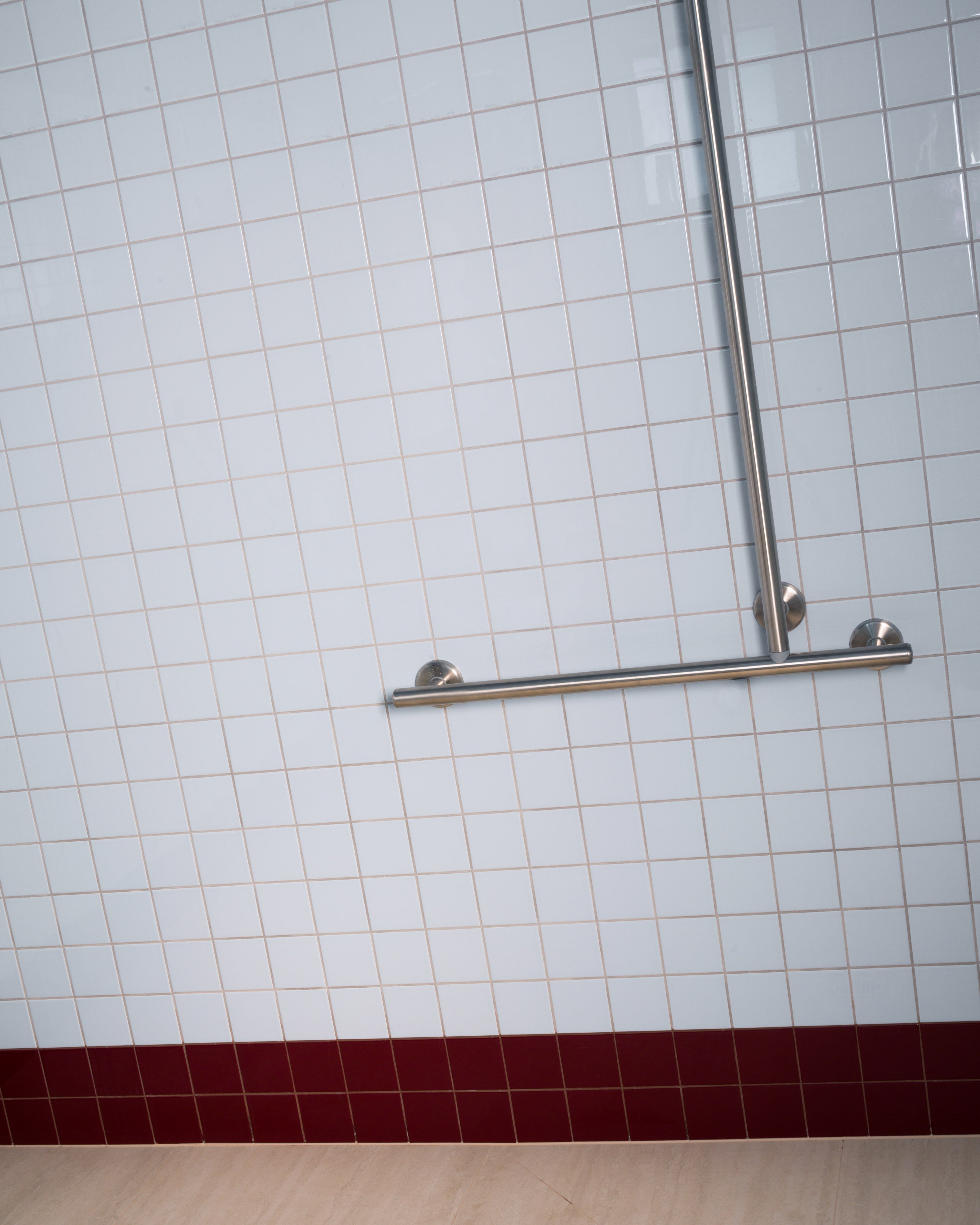 White square-tiled bathroom wall with a stainless steel grab rail, deep burgundy tile band, and timber flooring — accessible bathroom interior design detail by Studio Nicholas Noosa Queensland Australia