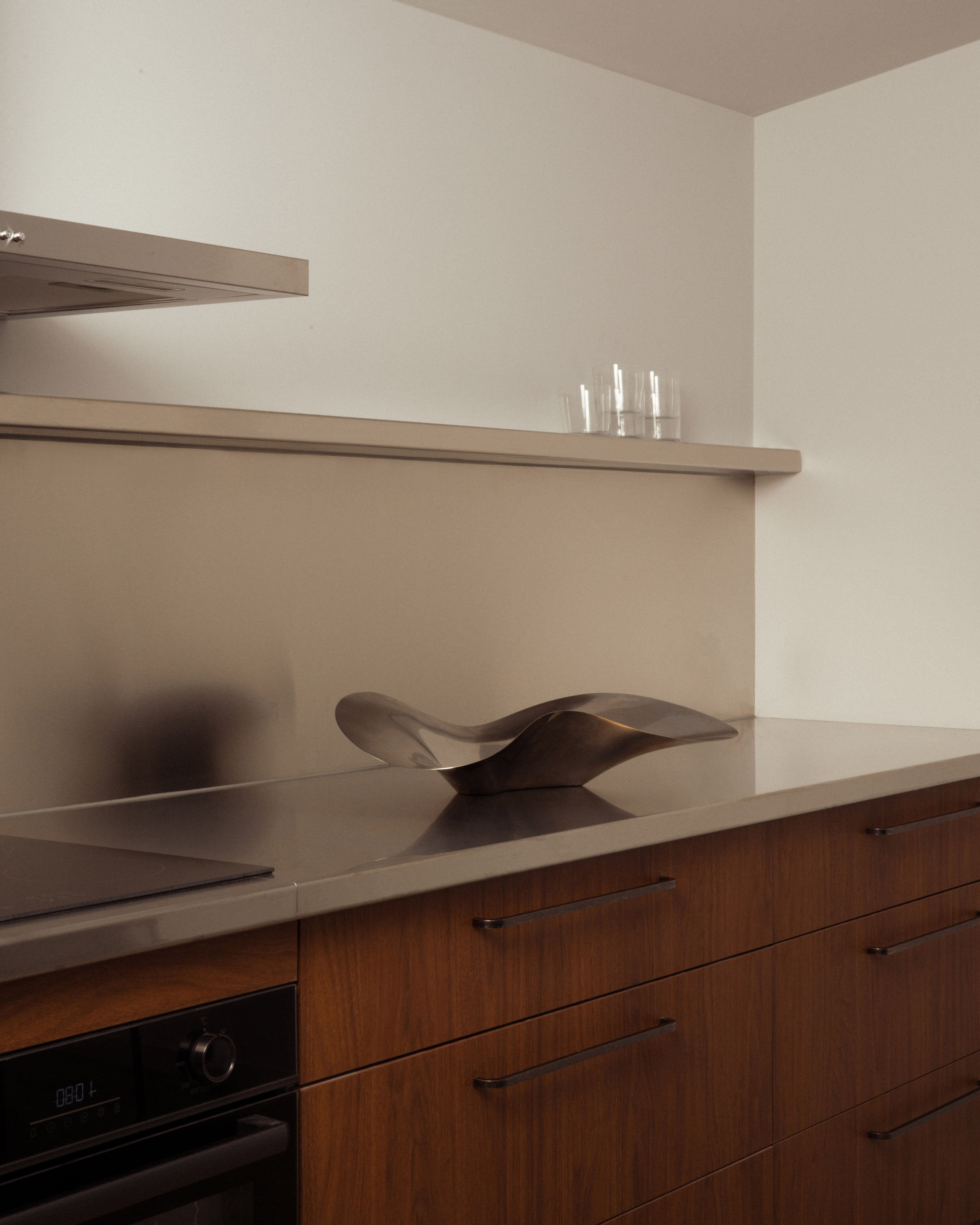Sculptural polished bronze abstract sculpture displayed on a stone kitchen benchtop with warm timber cabinetry and floating shelf styled with glassware — residential kitchen interior design styling by Studio Nicholas Noosa Queensland Australia