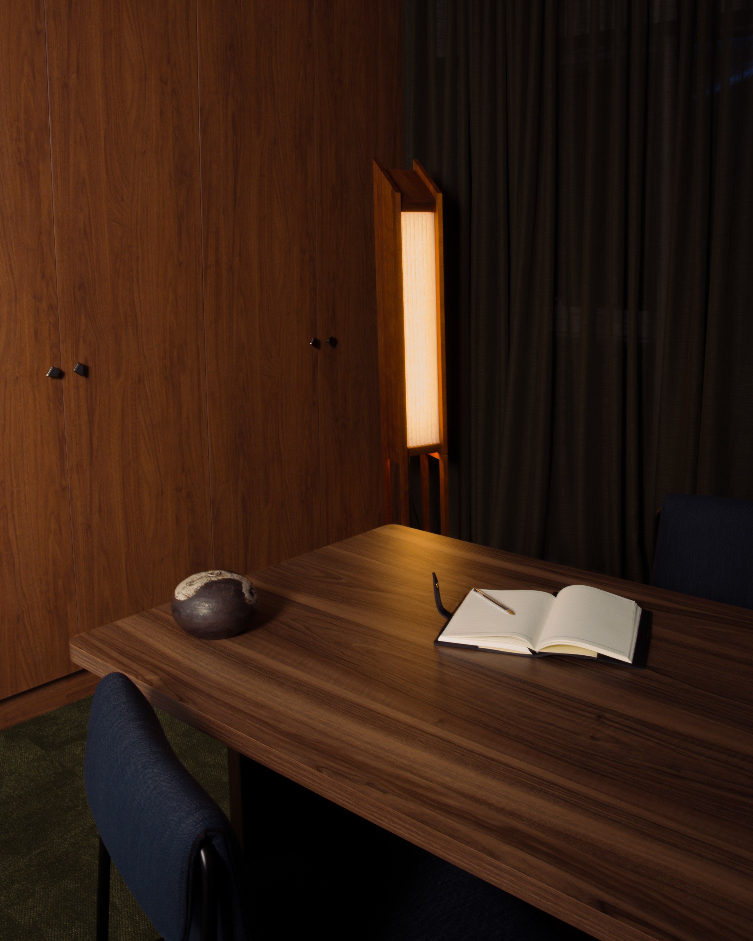 Warm timber-panelled meeting room with a natural timber table styled with an open book and dark ceramic teapot, dark upholstered chairs, and a glowing paper floor lamp beside drapery — commercial and hospitality interior design by Studio Nicholas Noosa Queensland Australia