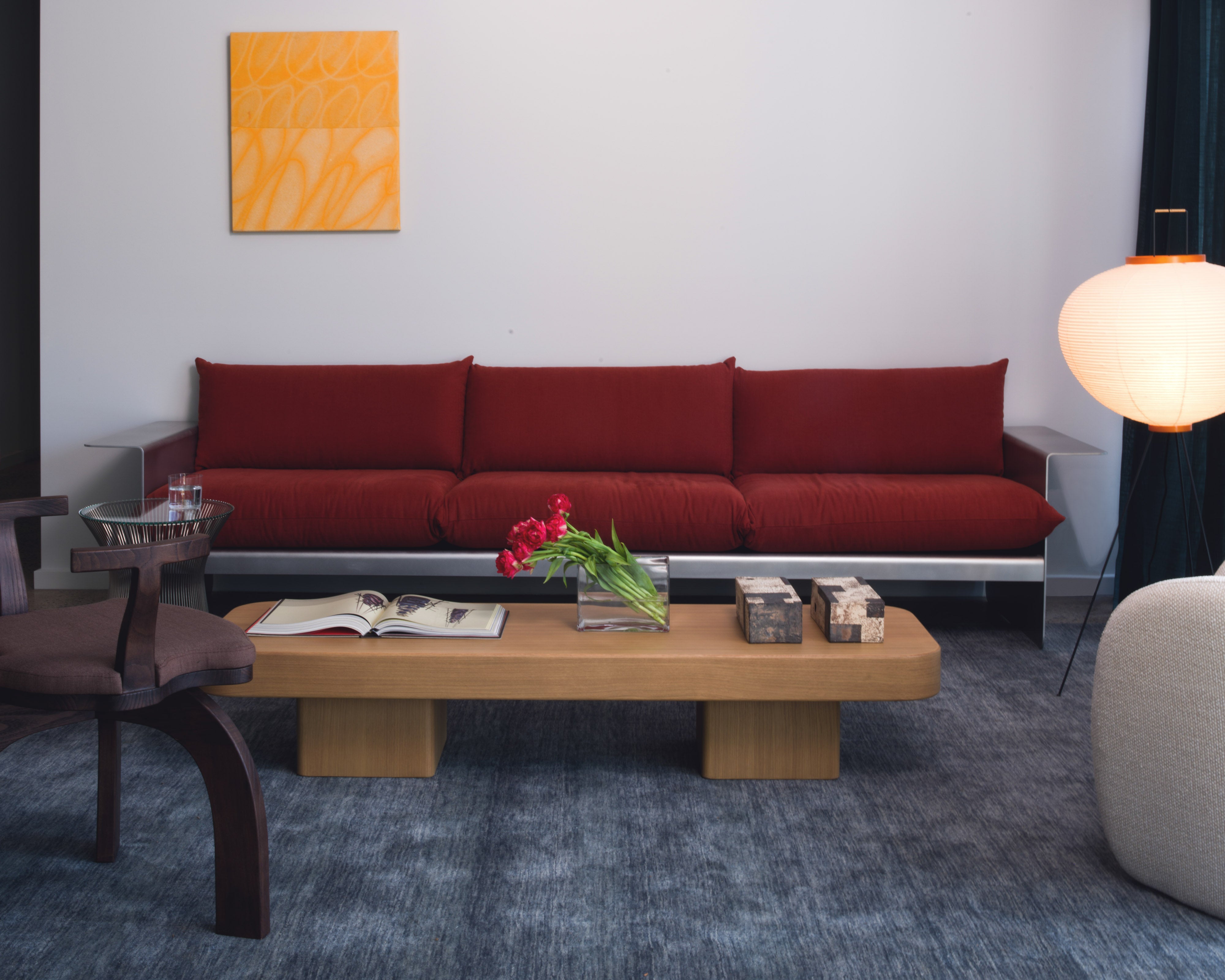 Contemporary residential living room with a red modular sofa, low natural oak coffee table styled with red tulips and an open book, orange abstract artwork, Akari paper globe floor lamp, and blue-grey wool carpet — luxury residential interior design by Studio Nicholas Noosa Queensland Australia